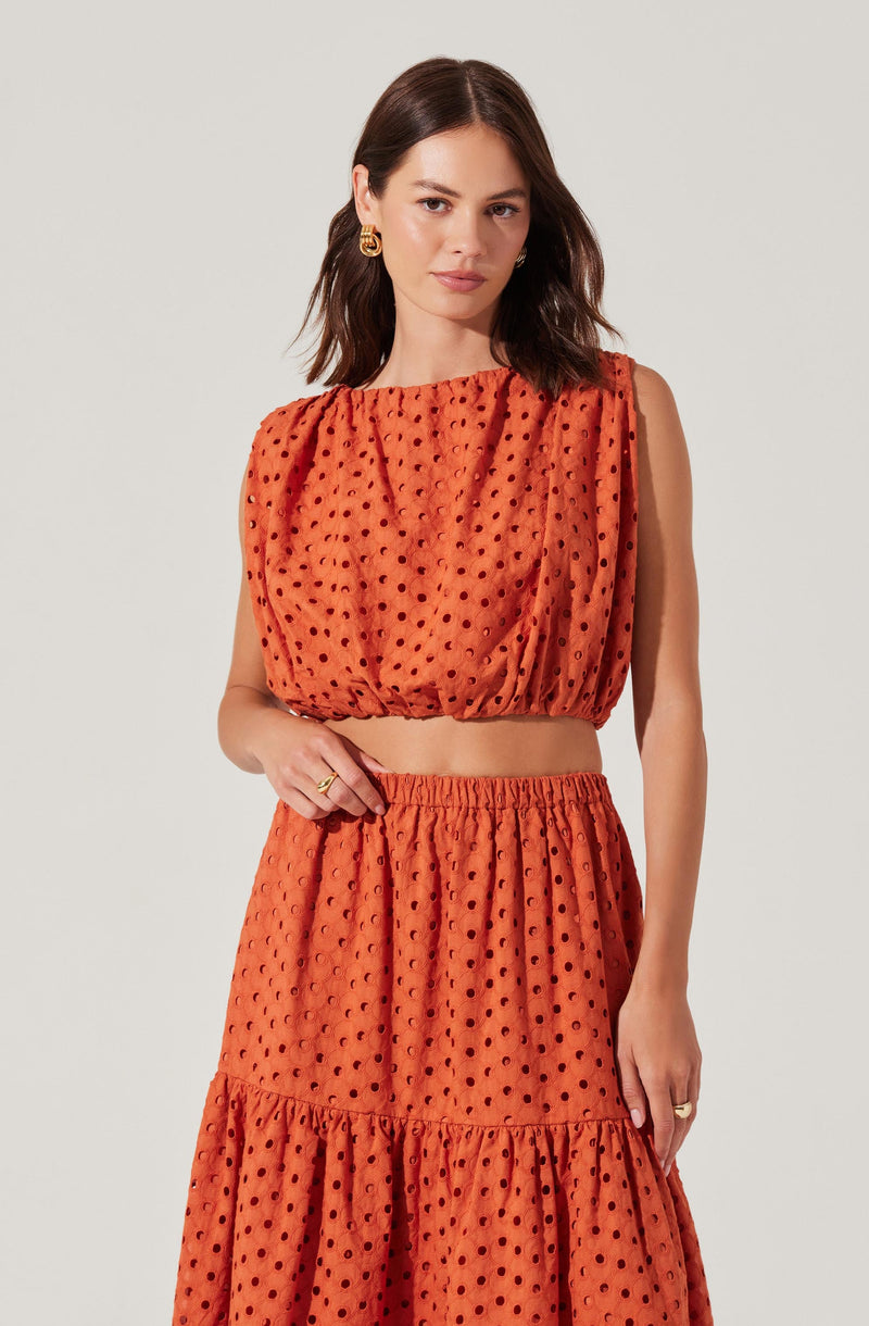 A woman is wearing a sleeveless, orange, dotted two-piece outfit consisting of a cropped top with a gathered neckline and a matching high-waisted, tiered skirt with a ruffled hem. The fabric has a textured appearance with evenly spaced black polka dots.