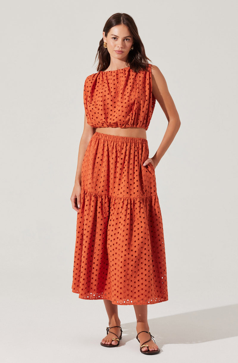 A woman is wearing a two-piece outfit made of orange fabric with a black polka dot pattern. The top is a sleeveless, loose-fitting crop top with a gathered neckline and armholes. The matching skirt is high-waisted with an elastic waistband, gathered at the waist, and has two tiers, creating a flowing, ankle-length skirt. She is also wearing black strappy sandals and gold hoop earrings.