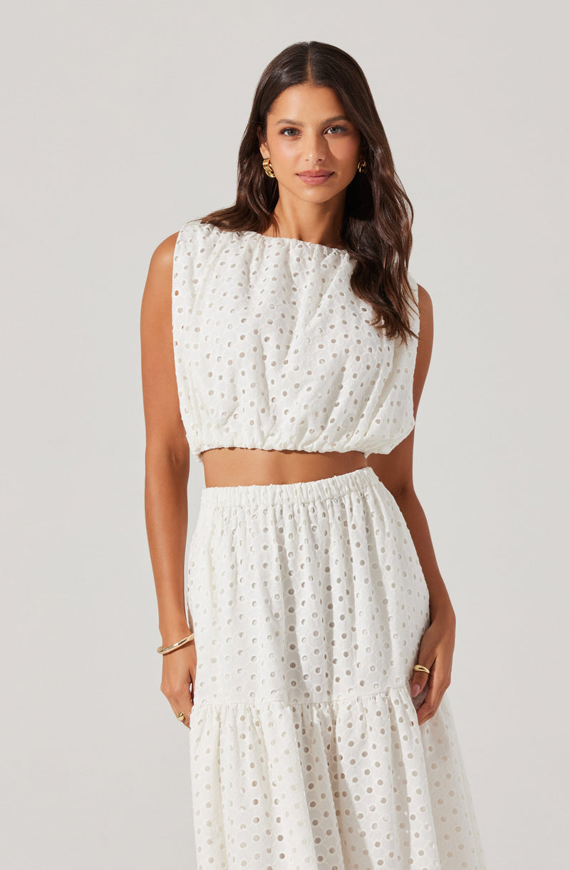 A woman is wearing a sleeveless, white, eyelet two-piece outfit. The top is cropped with a loose fit and gathered neckline, while the skirt is high-waisted with an elastic waistband, flowing into a tiered design. The fabric features a pattern of evenly spaced circular cutouts.