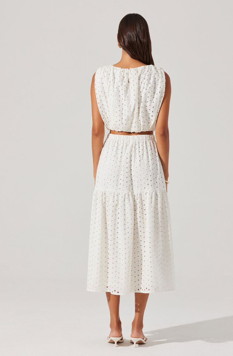 A woman is wearing a sleeveless white dress with an eyelet pattern. The dress has a loose, gathered bodice with a round neckline and a cropped waistline. The skirt is tiered, with a gathered seam at the mid-length and a flowing, ankle-length hem. She is also wearing white heeled shoes.