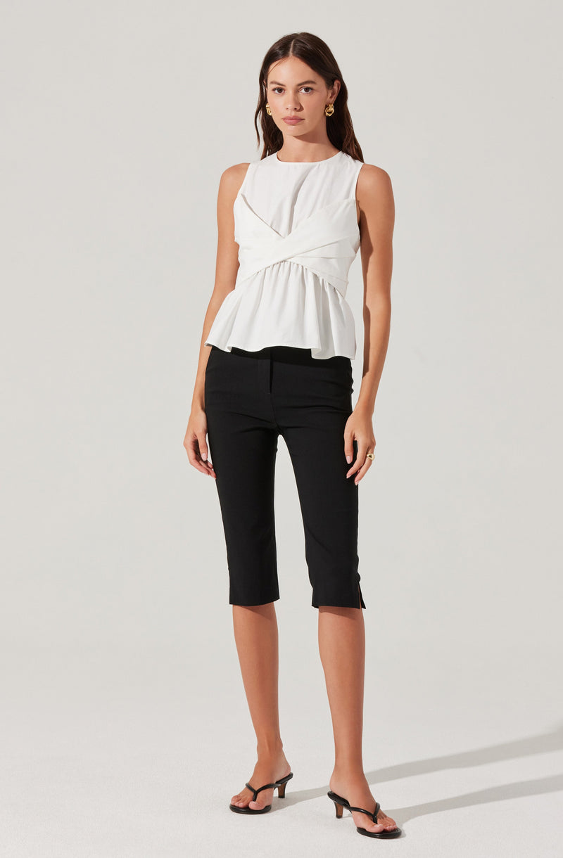 A woman is wearing a sleeveless white top with a draped, layered design and a ruffled hem, paired with black cropped pants that have small side slits at the hem. She is also wearing black open-toe high-heeled sandals and gold earrings.