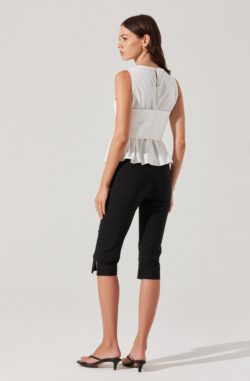 A woman is wearing a sleeveless, light-colored top with a fitted waist and a ruffled peplum hem. The top features a keyhole opening at the back. She pairs it with black cropped pants that have a small slit at the hem. She completes her outfit with black heeled sandals.