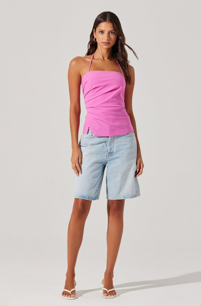 A woman is wearing a bright pink sleeveless halter top with side ruching details and small side slits, paired with light blue denim Bermuda shorts. She is also wearing white strappy high-heeled sandals.