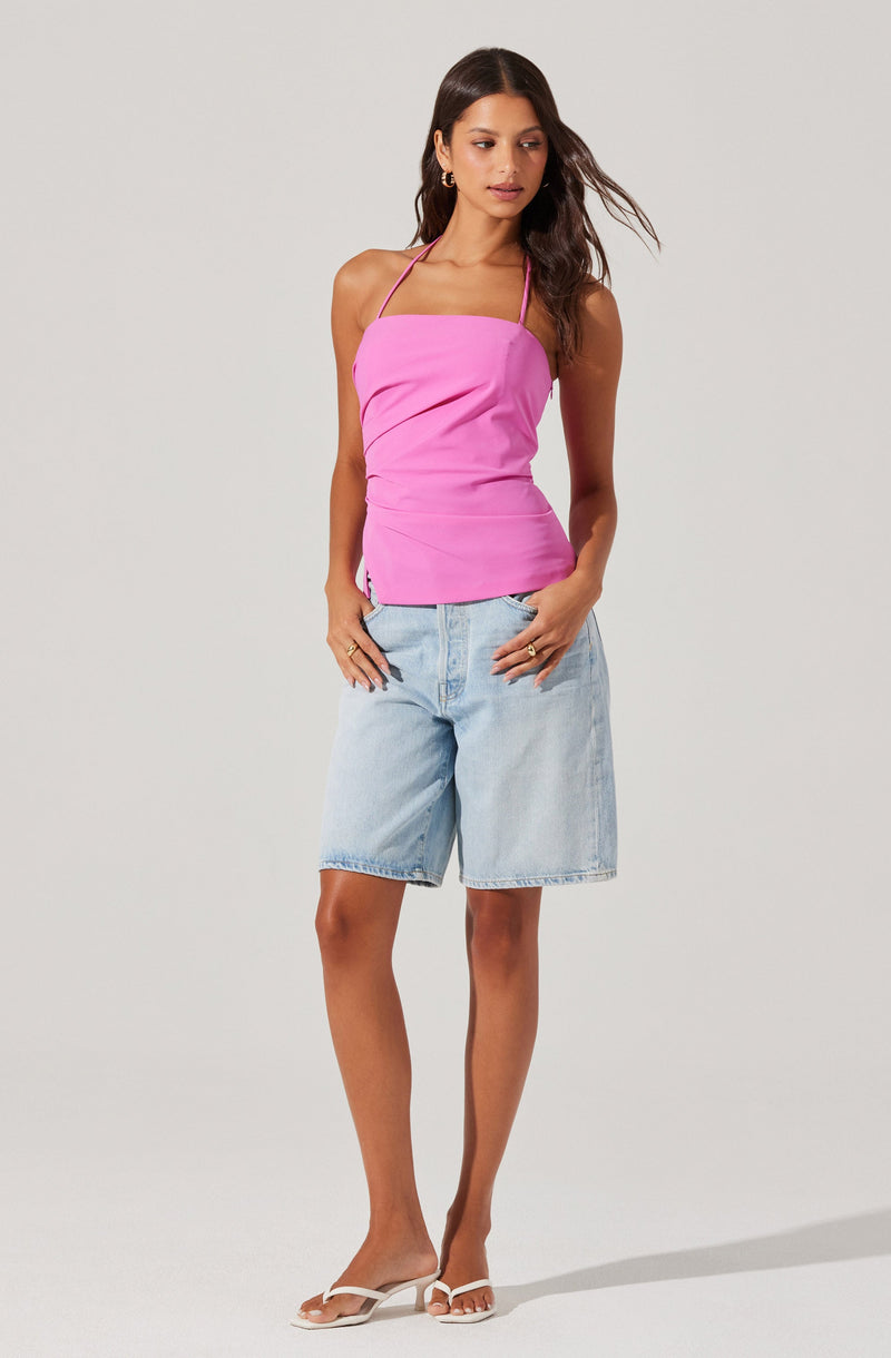 A woman is wearing a bright pink sleeveless top with thin spaghetti straps and a square neckline. She is also dressed in light blue denim knee-length shorts. She is wearing white open-toe sandals and gold hoop earrings.