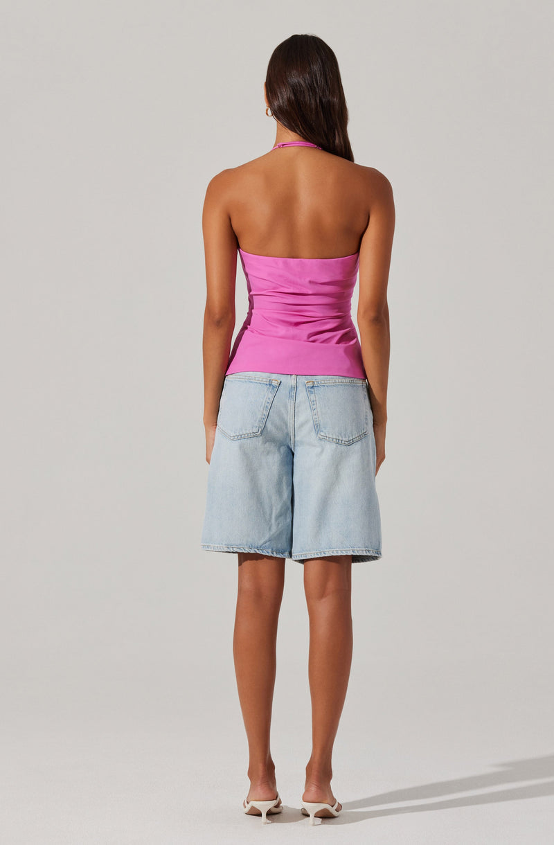 A woman is wearing a strapless, shiny pink top that is fitted and ruched around the waist. She is also wearing light-wash, knee-length denim shorts with large back pockets. She has on white high-heeled shoes.