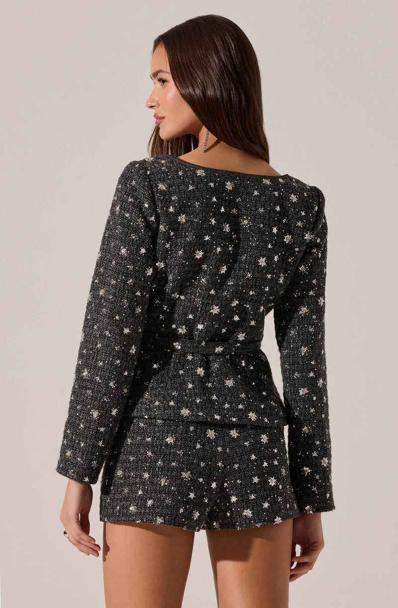 A woman is wearing a matching black tweed blazer and shorts set. The blazer has a textured, woven appearance with a pattern of small white stars and dots, long sleeves, and a fitted waist. The shorts are made from the same fabric and pattern, with a high waist and tailored fit.