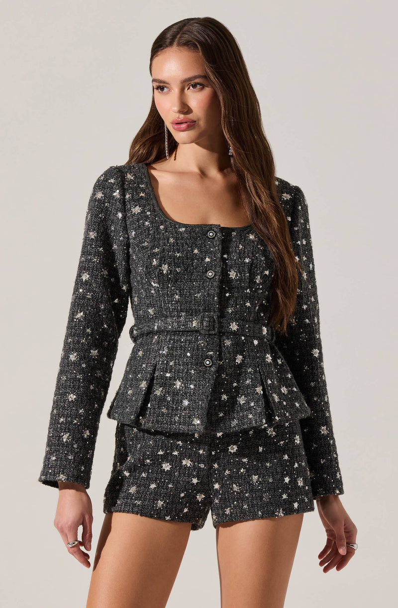 A woman is wearing a matching set of a black, sparkly tweed jacket and shorts. The jacket features long sleeves, a rounded neckline, and front buttons, with a textured, glittering star pattern throughout. The shorts are high-waisted and made from the same sparkly tweed fabric, coordinating with the jacket for a stylish, coordinated look.