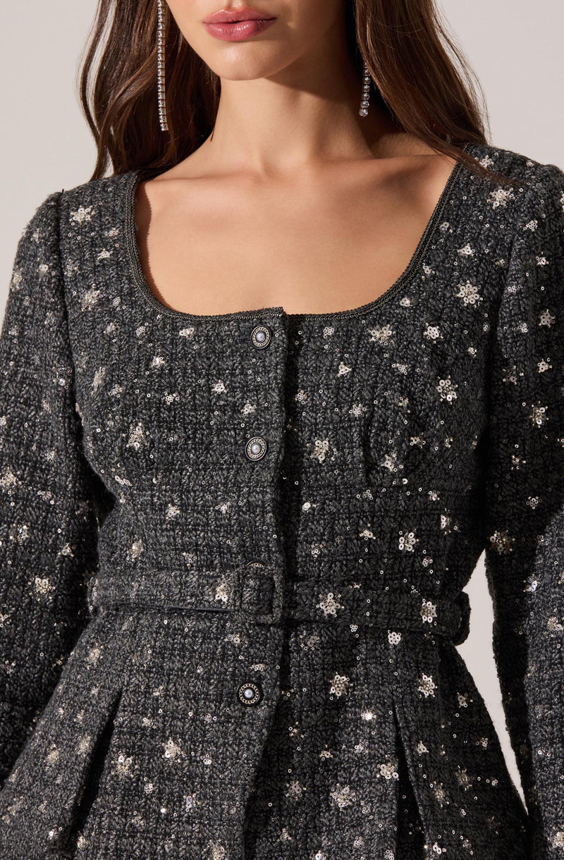 A woman is wearing a black, long-sleeved jacket adorned with silver sequins and floral embellishments. The jacket features a rounded neckline and is fastened with decorative buttons down the front. The fabric is textured with a glittery, shiny appearance.