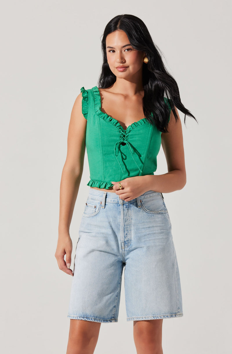A woman is wearing a sleeveless green top with ruffled shoulder straps and a front drawstring detail. She is also wearing light-wash, high-waisted denim shorts with a relaxed fit.