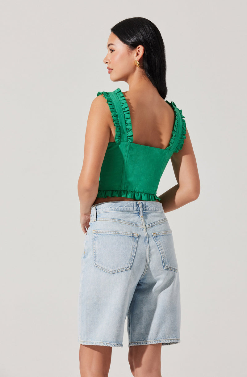 A woman is wearing a sleeveless green top with ruffled straps and a ruffled hem, paired with light-wash denim shorts.