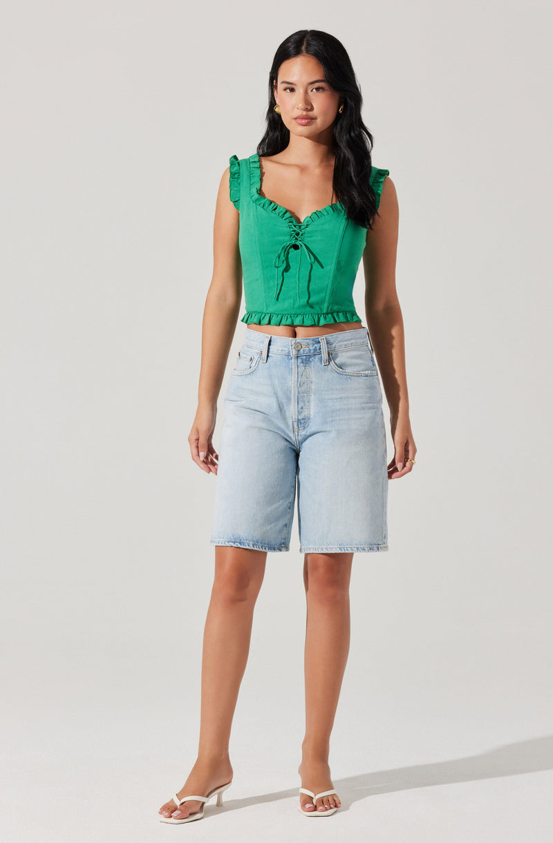 A woman is wearing a sleeveless, bright green crop top with ruffled shoulder straps, a ruched front with a tie detail, and a ruffled hem. She pairs it with light-wash denim Bermuda shorts and white strappy high-heeled sandals.