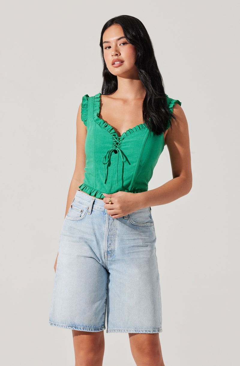 A woman is wearing a bright green sleeveless top with ruffled shoulder straps and a gathered neckline with a lace-up detail in the front. The top has a fitted style with an elasticized hem. She is also wearing high-waisted, light-wash denim shorts with a relaxed fit.