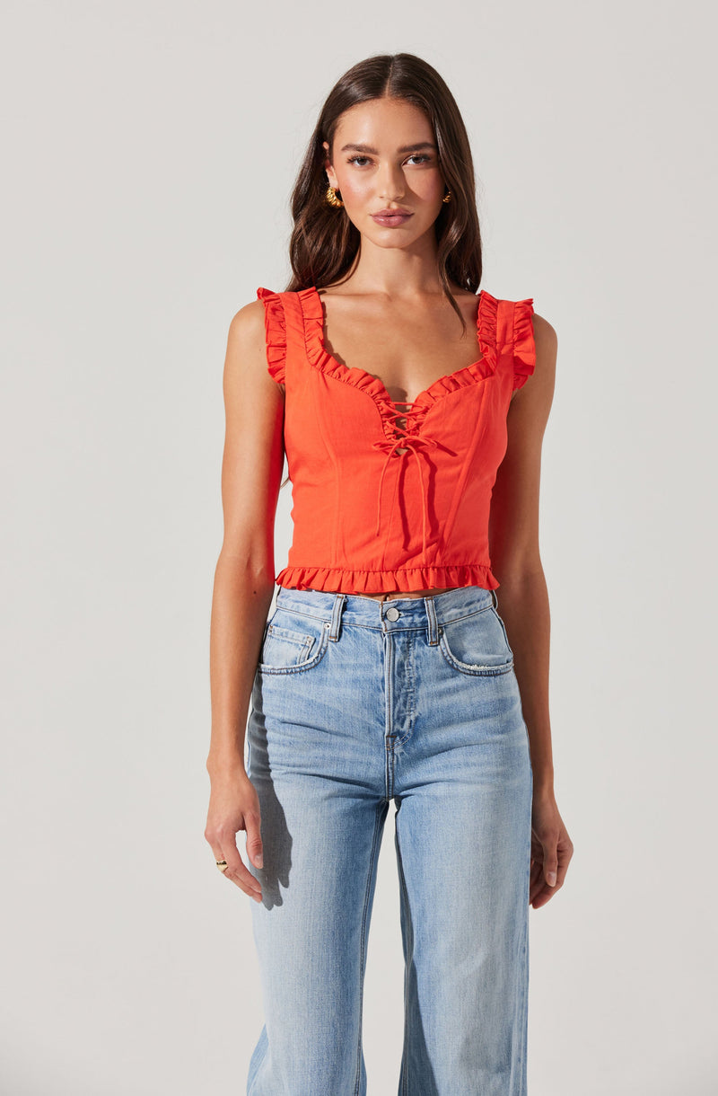 A young woman is wearing a bright red sleeveless crop top with ruffled shoulder straps and a lace-up front detail. She pairs it with high-waisted, light-wash blue jeans. The top has a slightly ruffled hemline.