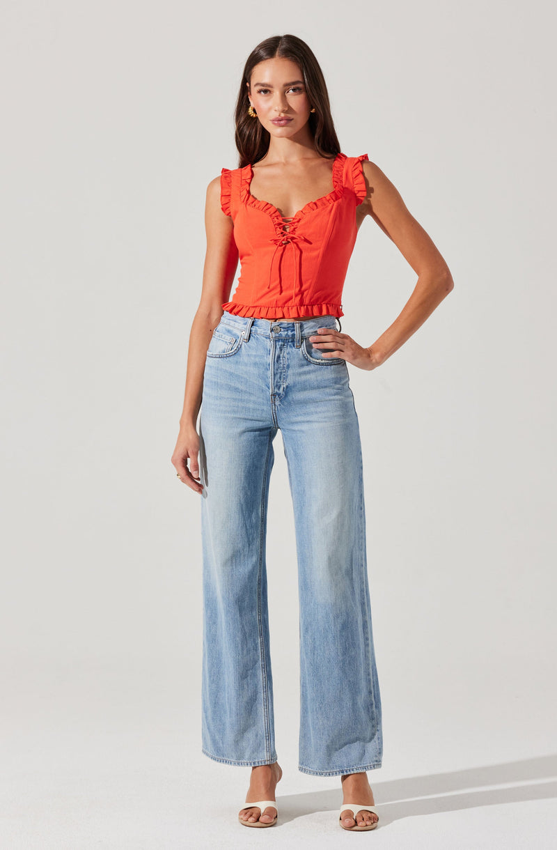 A woman is wearing a sleeveless, bright red top with ruffle details on the shoulders and a lace-up front. She is also wearing light-wash, high-waisted, wide-leg jeans.