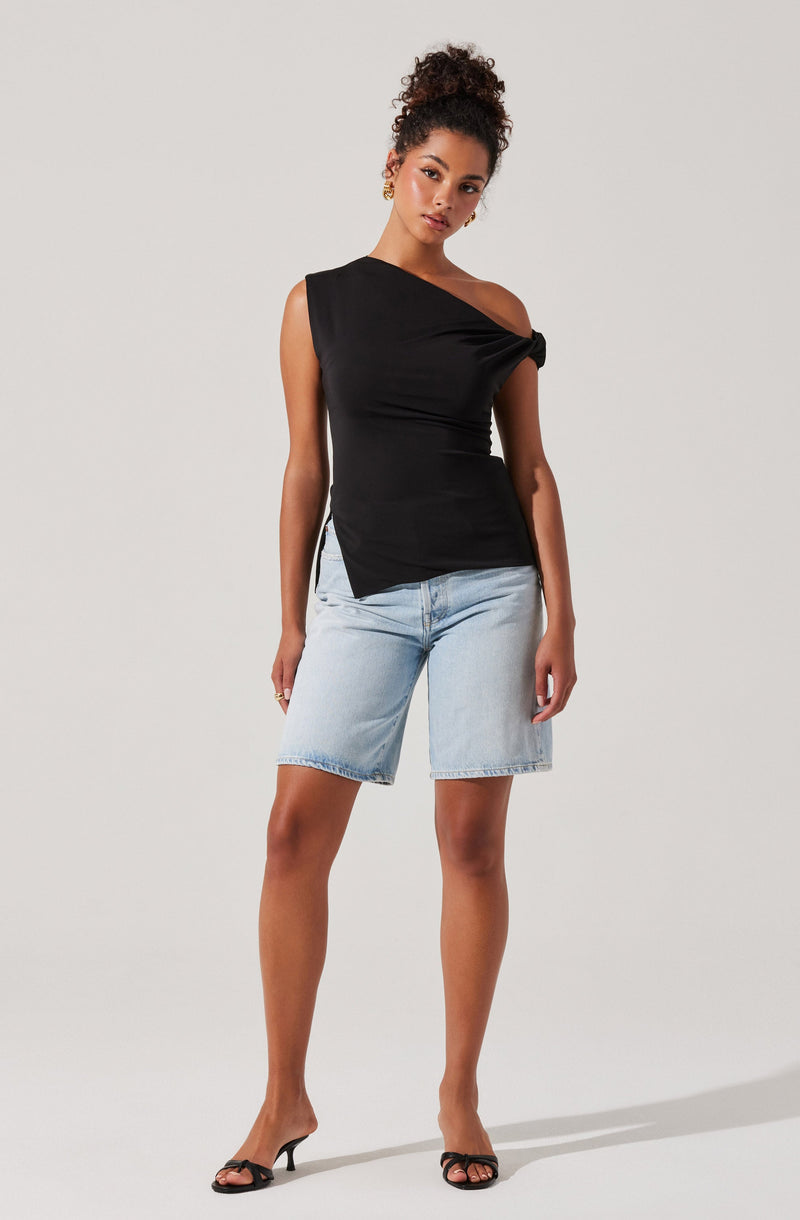 A woman is wearing a black sleeveless top with an asymmetrical neckline and a knot detail on one shoulder. She has paired it with light-wash, knee-length denim shorts and black open-toe high heels.