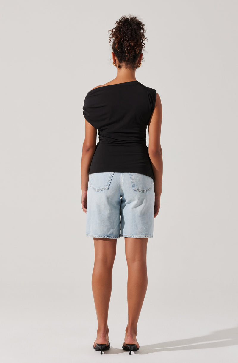 A woman is wearing a black sleeveless top with a one-shoulder design, featuring ruching detail on the shoulder, paired with light-wash, knee-length denim shorts with rolled-up hems and back pockets. She is also wearing open-toe heels.