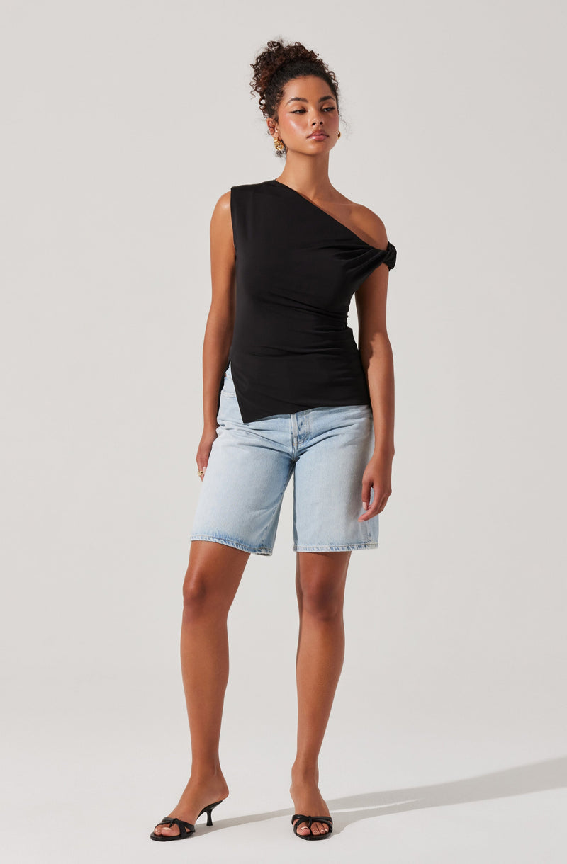 A woman is wearing a black, one-shoulder top with a twist detail at the shoulder, combined with light blue denim knee-length shorts. She has accessorized with hoop earrings and is wearing black high-heeled sandals.