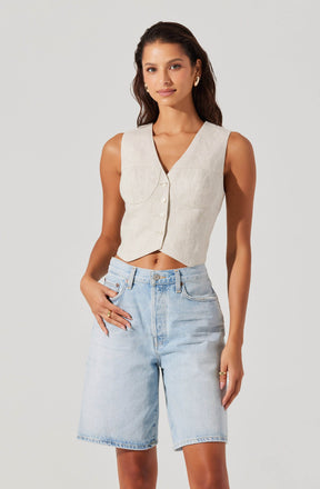 A woman is wearing a sleeveless, beige crop top with a V neckline and front buttons. She has paired it with high-waisted, light blue denim shorts that reach just above the knees, with a relaxed fit.
