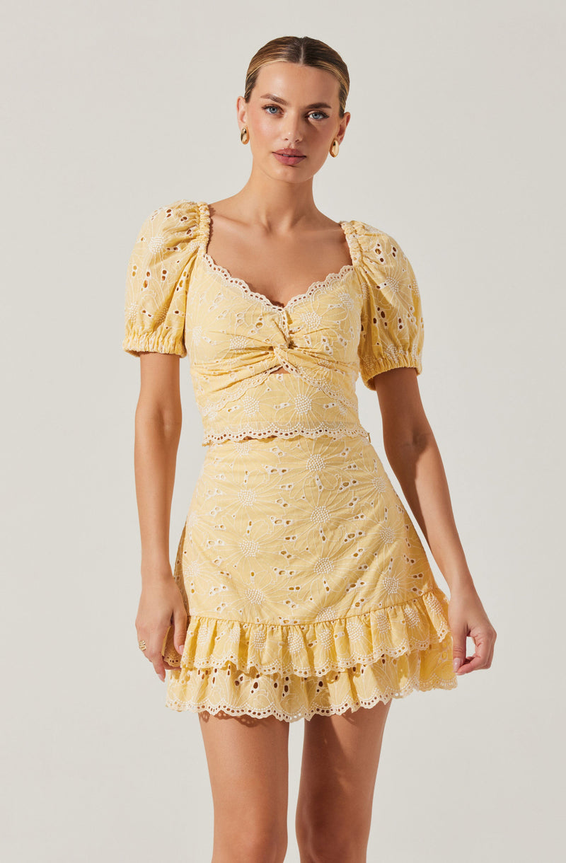 A woman is wearing a matching yellow outfit consisting of a top with short, puffed sleeves and a ruffled hem, both made from fabric with eyelet embroidery and floral patterns. The top features a scalloped neckline and a knot detail at the front. She is also wearing a high-waisted skirt with the same embroidered fabric, decorated with ruffles at the hem.