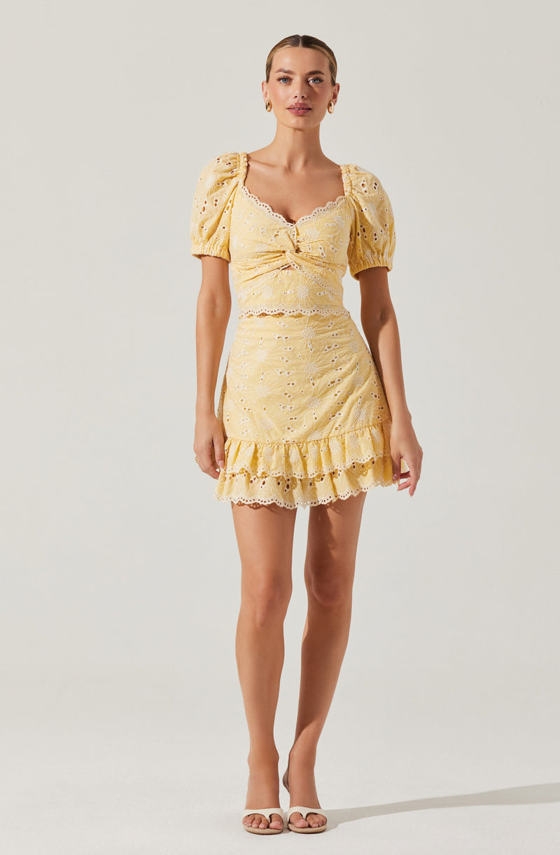 A woman is wearing a yellow eyelet lace dress with puffed short sleeves and a fitted bodice that features a small bow detail at the center. The skirt is short with a ruffled hem, and the eyelet design creates a textured, openwork pattern throughout the dress. She is also wearing open-toe high-heeled sandals.