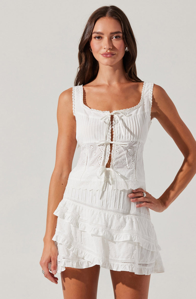 A woman is wearing a sleeveless white dress with lace and eyelet details. The top has a square neckline with lace trim and a front tie closure. The skirt features layered ruffles with eyelet embroidery.