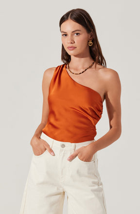 A woman is wearing a sleeveless, one-shoulder top made of shiny, burnt orange satin fabric. She has paired it with high-waisted, off-white pants. She accessorized with gold hoop earrings and a beaded necklace.