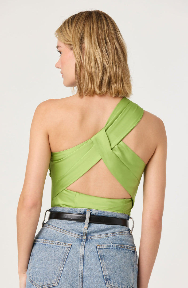 A woman is wearing a lime green satin one-shoulder top with a crisscross design on the back, featuring cut-out sections. She pairs the top with high-waisted, light-wash jeans and a black belt.