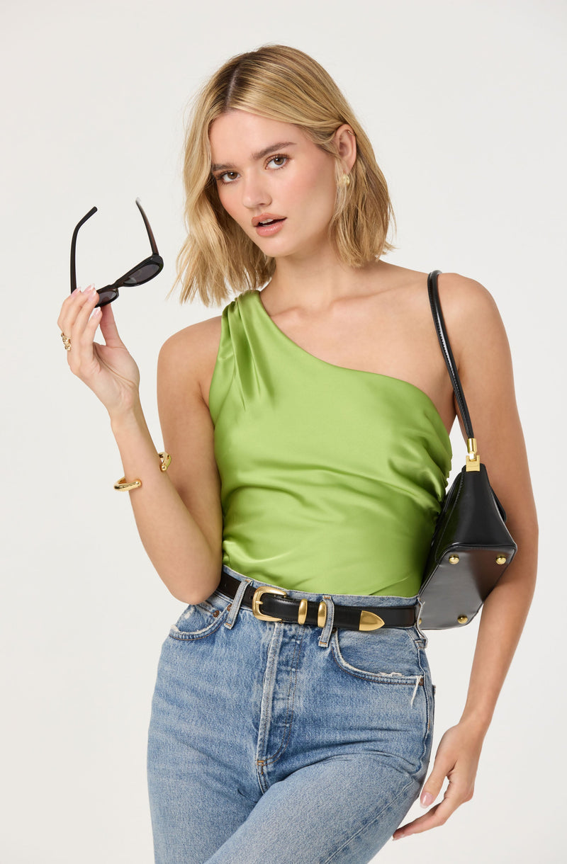 A woman is wearing a bright green satin one-shoulder top, paired with high-waisted light blue jeans. She has on a black belt with a gold buckle, and is carrying a small black handbag with gold accents on her right shoulder. She is holding a pair of black sunglasses in her right hand, and her left hand rests on her hip.