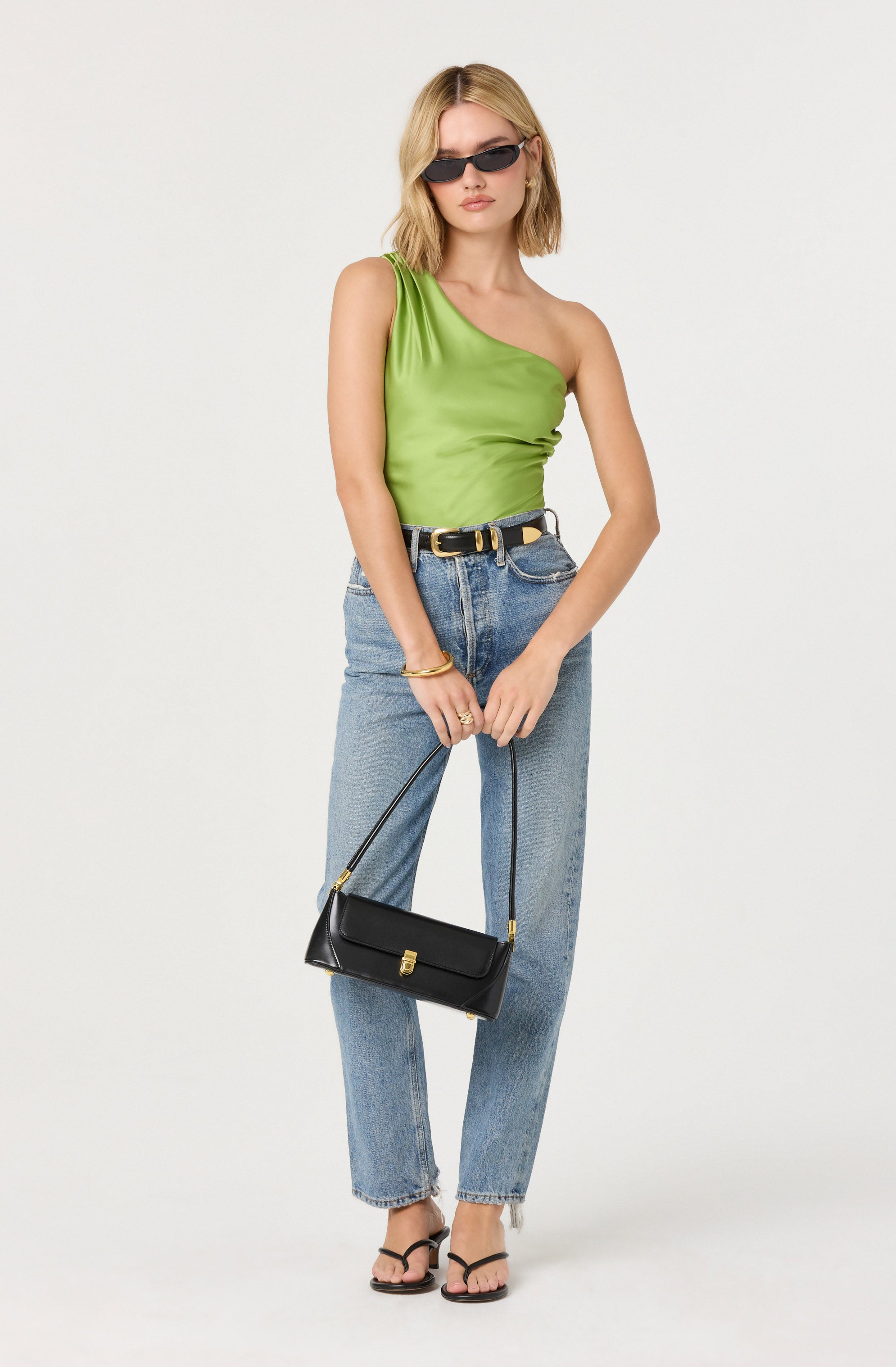 A woman is wearing a bright green, one-shoulder satin top paired with high-waisted, light-wash jeans. She accessorizes with black sunglasses, a black belt with a gold buckle, and gold jewelry. She is holding a black handbag with a gold clasp and is wearing black strappy sandals.