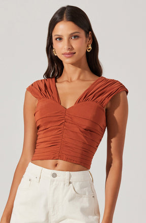 A woman is wearing a rust-colored, ruched crop top with gathered fabric details on the shoulders and across the bust. She is also wearing high-waisted, off-white pants.