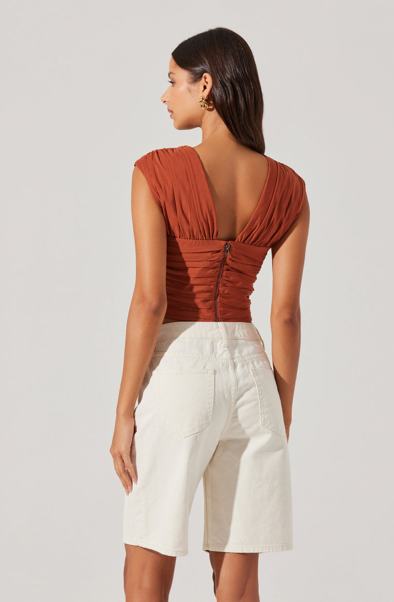 A woman is wearing a rust-colored, ruched, sleeveless top with a deep V back and a zipper closure running down the center back. She pairs it with high-waisted, off-white Bermuda-length shorts. The top features gathered fabric details on the shoulders and around the waist.