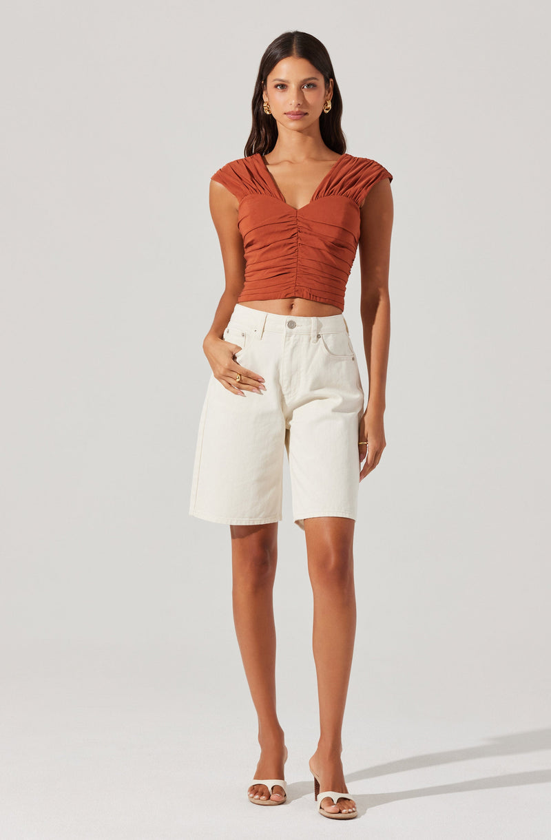 A woman is wearing a fitted, rust-colored crop top with ruched detailing and capped sleeves, paired with high-waisted, knee-length white shorts. She is also wearing open-toe heels and gold jewelry.