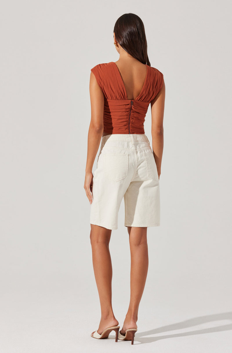 A woman is wearing a rust-colored, pleated top with cap sleeves and a deep V-back, secured with a zipper at the middle of the back. She is also wearing high-waisted, knee-length white shorts with a relaxed fit and large front pockets. She is wearing beige high-heeled sandals.