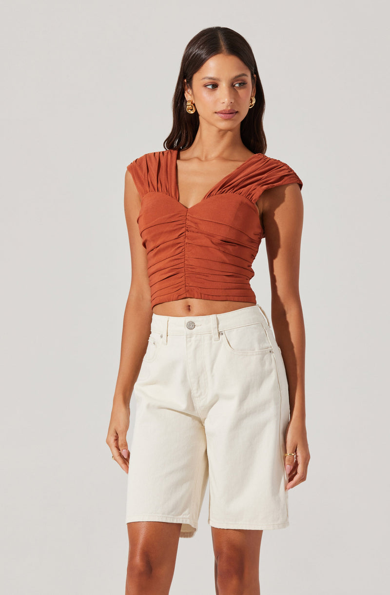 A woman is wearing a ruched, rust-colored sleeveless crop top with gathered fabric around the shoulders and bust area. She has paired it with high-waisted, off-white knee-length shorts.