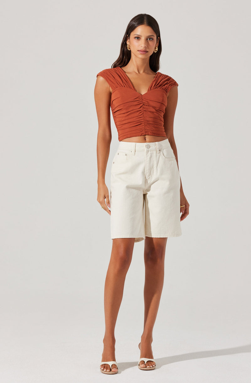 A woman is wearing a rust-colored, ruched, sleeveless crop top with gathered fabric and a V-neckline. She pairs it with high-waisted, white knee-length shorts. She is also wearing open-toed, beige high heels.