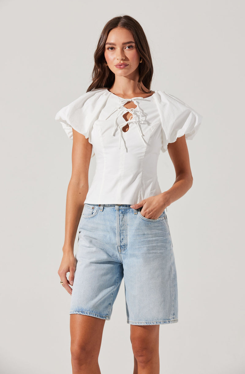 A woman is wearing a white top with puffed short sleeves and a lace-up front with tie closures, paired with light blue denim shorts.