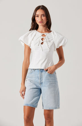 A woman is wearing a white top with puffed short sleeves and a lace-up front with tie closures, paired with light blue denim shorts.