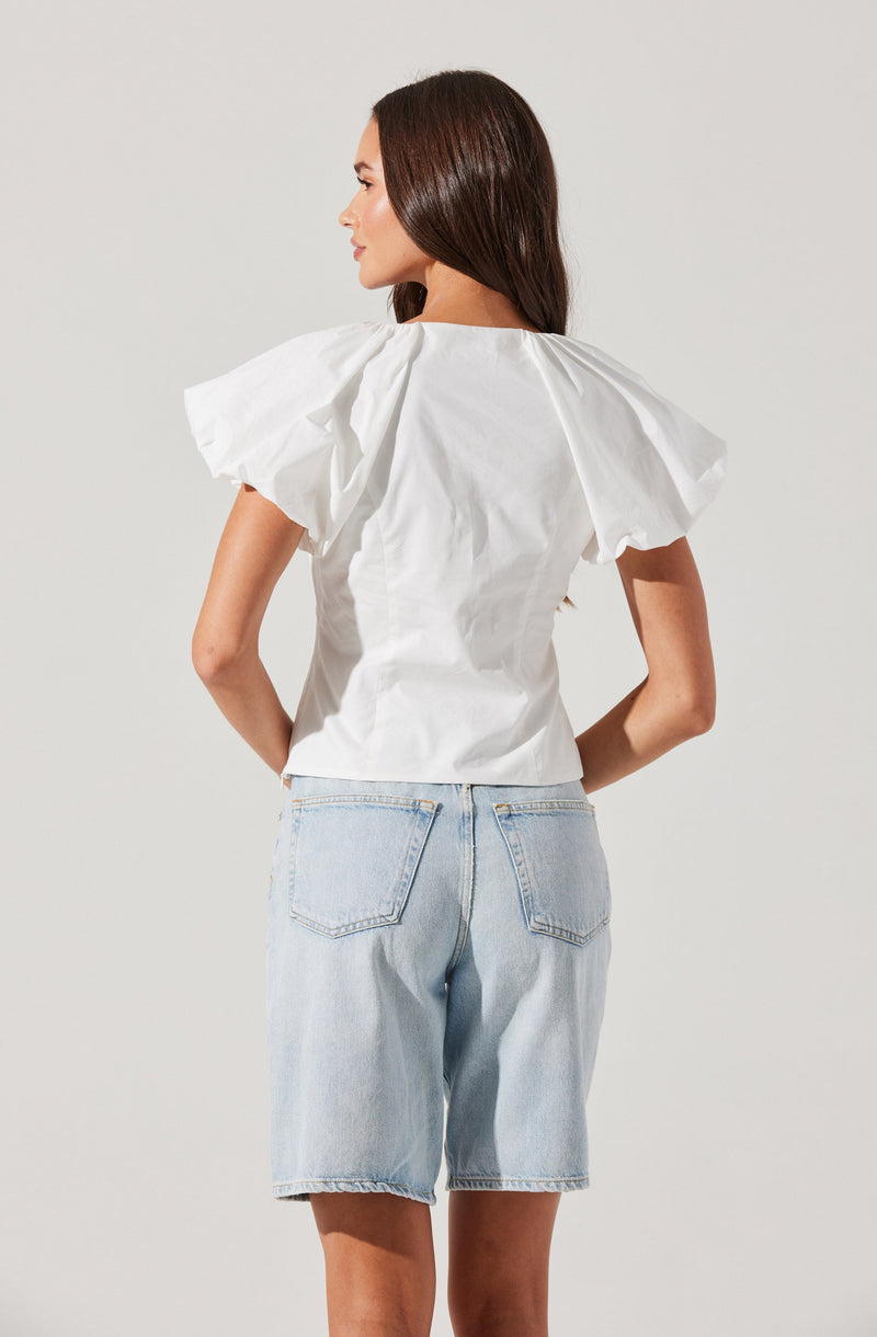 A woman is wearing a white blouse with large, puffed short sleeves and a fitted waist. She is also wearing light blue denim shorts that reach above her knees, with visible back pockets and a slightly frayed hem.