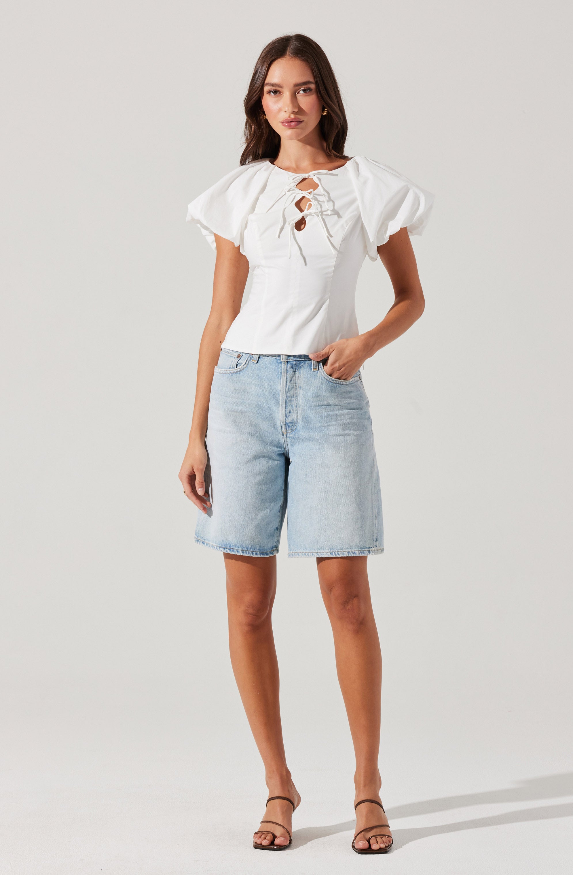 A woman is wearing a white top with puffed short sleeves and a front lace-up detail with cutouts. She is also wearing light-wash denim shorts that reach just above her knees. She has on a pair of open-toed, ankle-strap heels.