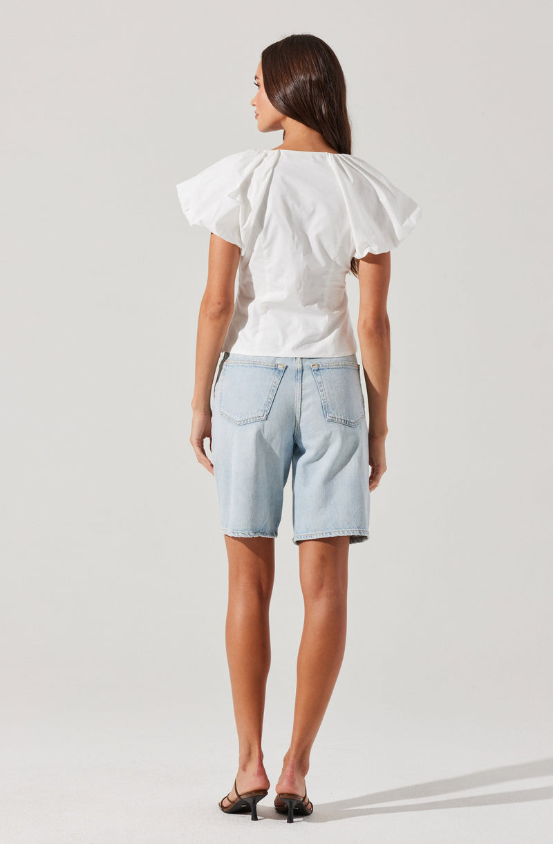 A woman is wearing a white top with large, puffed short sleeves and a round neckline. She pairs it with light-wash, knee-length denim shorts that have visible back pockets. She completes her outfit with black high-heeled sandals.