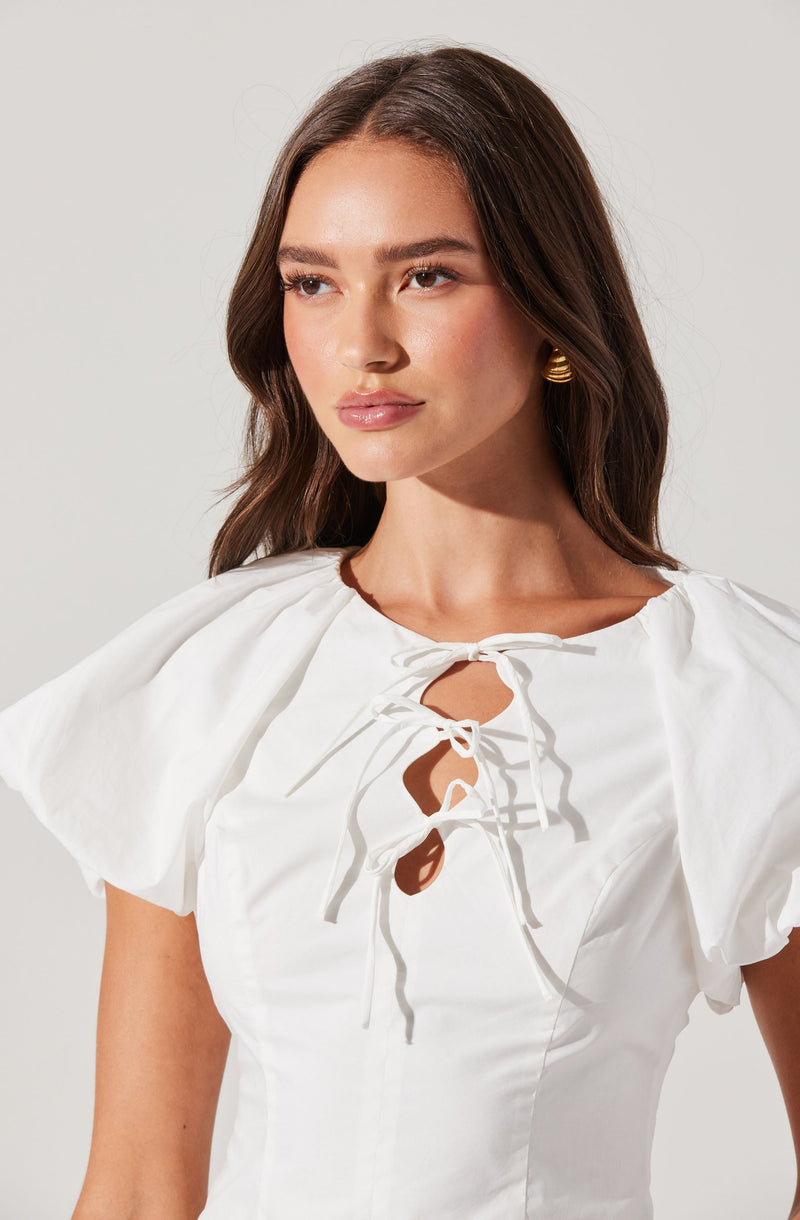 A woman is wearing a white dress with short puffed sleeves and a rounded neckline. The dress features a keyhole opening at the front, which is laced up with a drawstring tied into a bow, creating a decorative effect. She accessorizes with gold earrings.