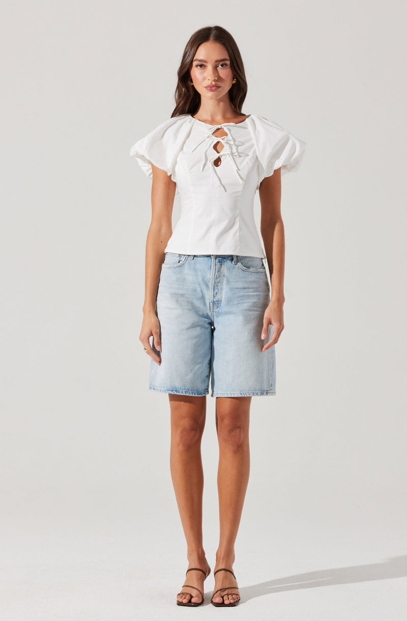 A woman is wearing a white short-sleeved blouse with puffed sleeves and a lace-up front detail with decorative cutouts. She pairs the top with light-wash, knee-length denim shorts and black strappy heeled sandals.