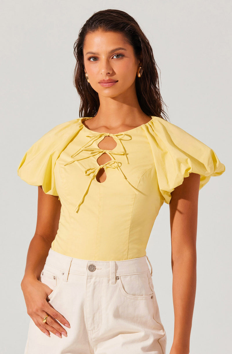 A woman is wearing a yellow blouse with puffed short sleeves and a round neckline. The blouse features three front keyhole cutouts, each tied with a matching yellow string. She is also wearing high-waisted, light-colored pants.