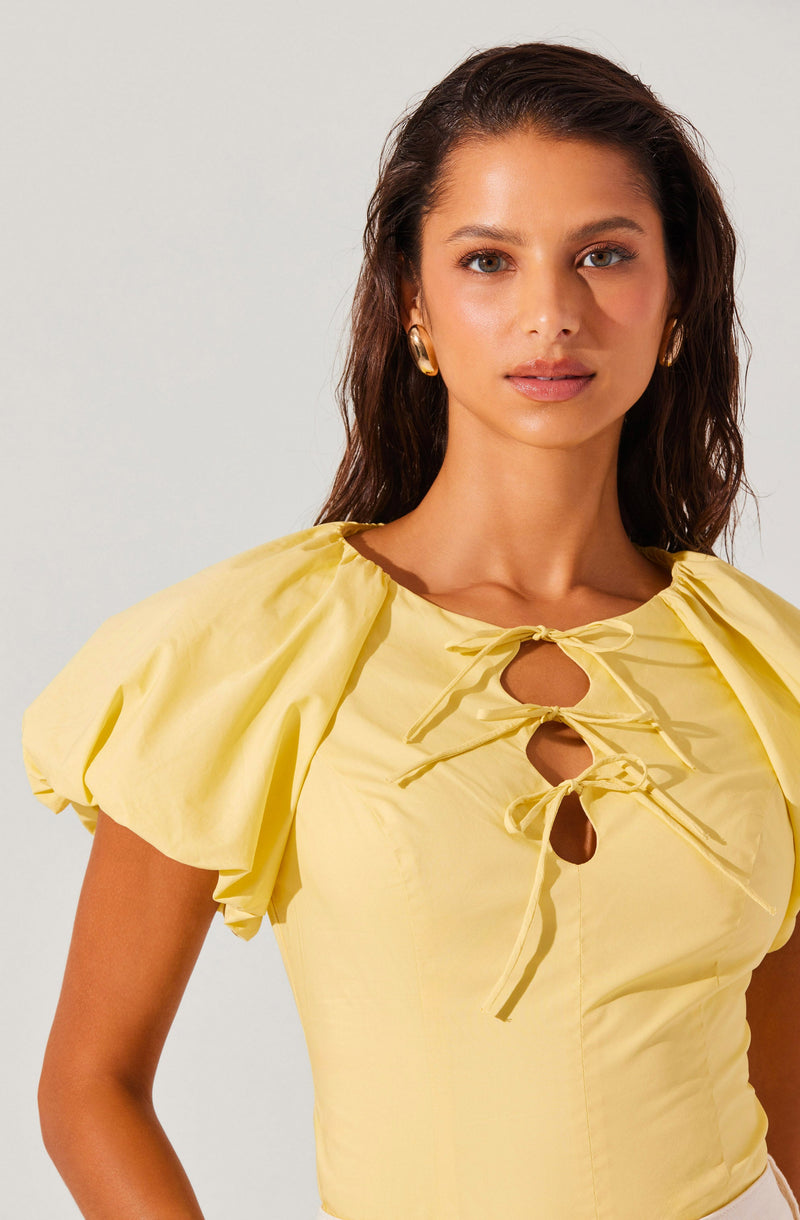 A woman is wearing a light yellow top with short, puffed sleeves. The top features a gathered neckline with three decorative bows threaded through keyhole cutouts at the front.