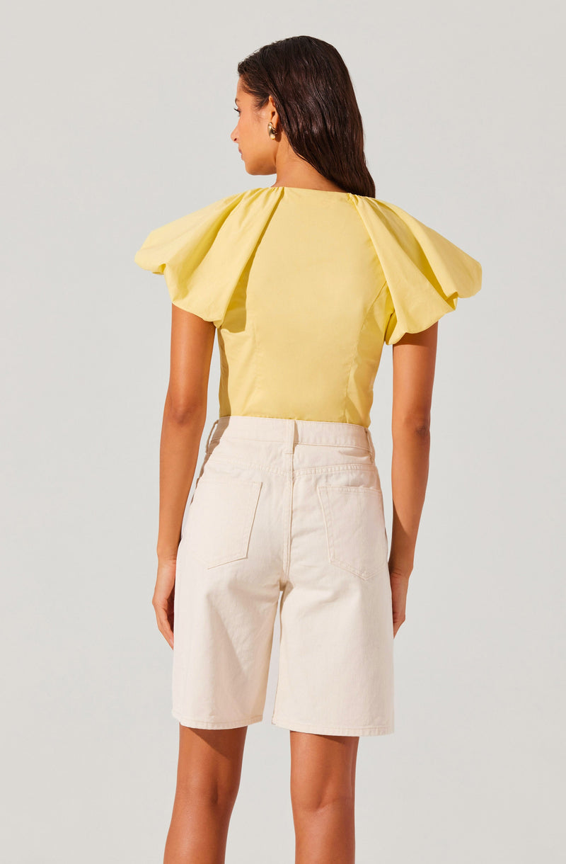 A woman is wearing a yellow top with oversized, puffed short sleeves that have a gathered detail at the shoulder. The top is fitted at the waist and made of a smooth, lightweight fabric. She pairs it with high-waisted, knee-length off-white shorts that have large back pockets and a relaxed fit.