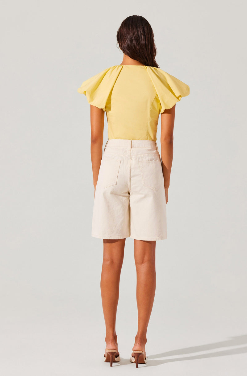 The image shows a woman wearing a yellow top with large, puffed short sleeves and a fitted bodice. She is also dressed in high-waisted, knee-length cream-colored shorts with visible stitching details. She has on beige high-heeled shoes. The woman has long, dark brown hair and is standing with her back to the camera.