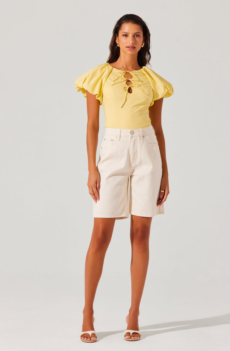 A woman is wearing a yellow top with puffed short sleeves and a lace-up front featuring circular cutouts. She pairs the top with high-waisted, knee-length white shorts that have front pockets. She completes the outfit with white open-toe sandals and minimal jewelry, including small earrings and rings.