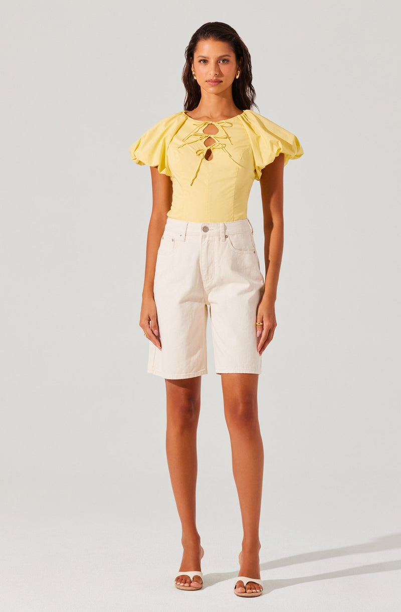 A woman is wearing a yellow short-sleeved top with puffed sleeves and front lace-up detailing with metal eyelets. She pairs it with high-waisted, knee-length white shorts and beige open-toe heeled sandals.
