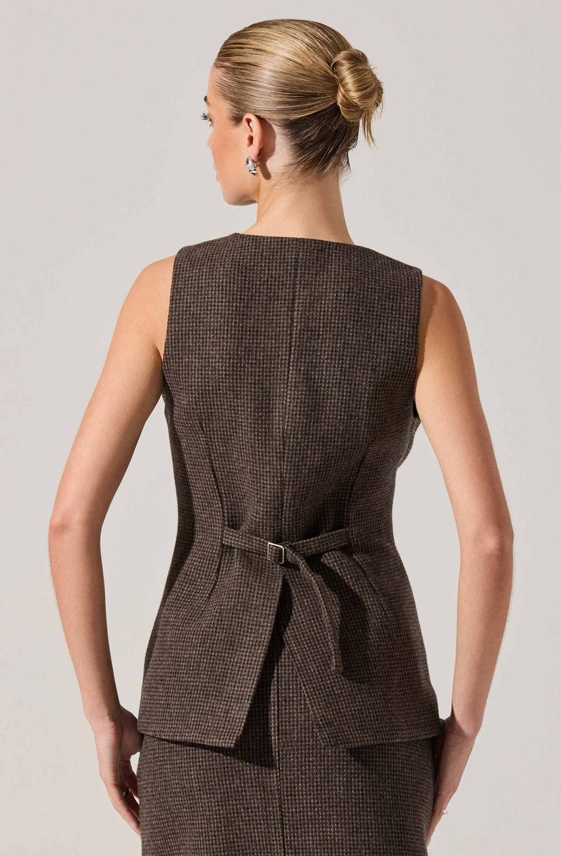 A woman is wearing a sleeveless brown houndstooth patterned blazer with a waist belt. The blazer has a fitted silhouette and a structured design, with a slight flare at the bottom. She has her hair styled in a neat, low bun and is accessorizing with earrings.