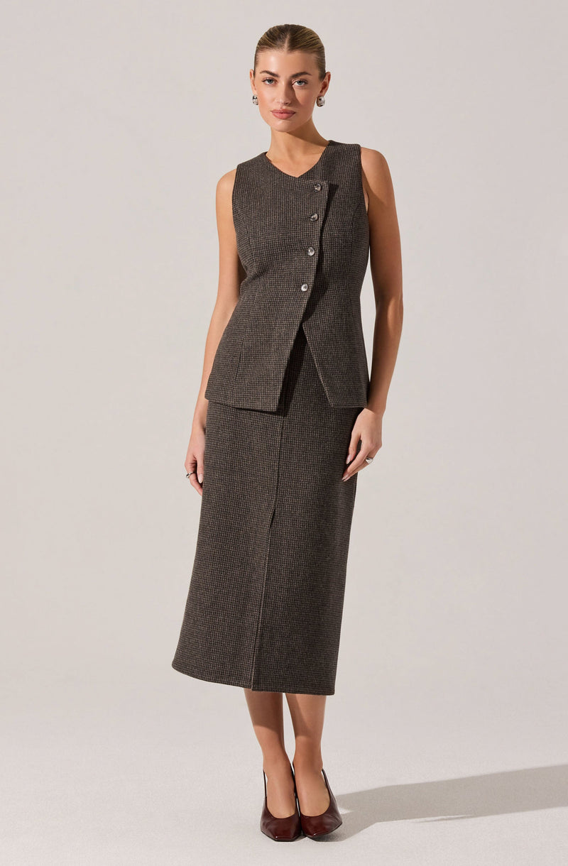 A woman is wearing a sleeveless, dark plaid tailored vest with a row of four buttons on the front and a matching long, fitted skirt that extends to her mid-calf. She is also wearing dark heels.